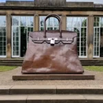 Large bronze handbag sculpture displayed outdoors on a pedestal in front of a building with tall glass windows.
