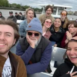A group of people sit closely together on an outdoor boat ride, with a city shoreline and cloudy sky in the background.