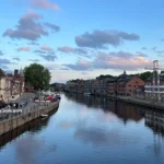 Riverside view of historic buildings, boats, and walkways along calm water under a blue sky with scattered clouds.