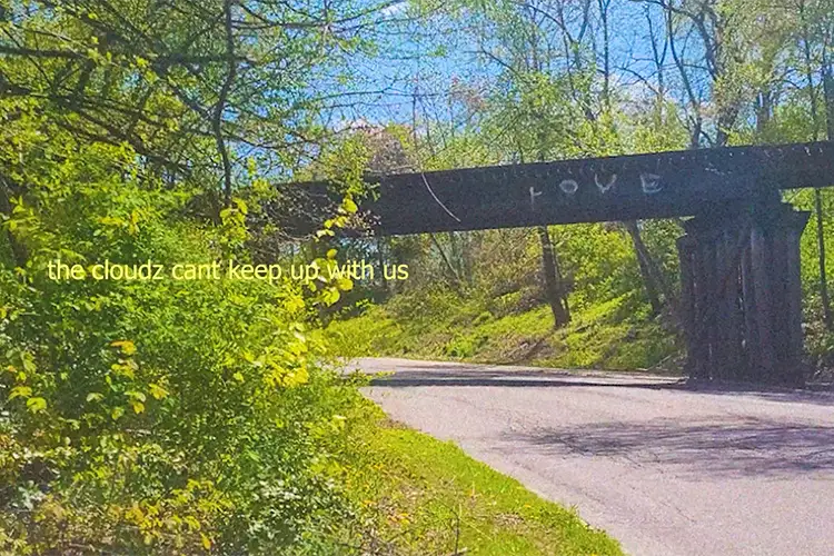 Road curving under a railroad bridge on a sunny day. Faint text over the image reads "the cloudz cant keep up with us".