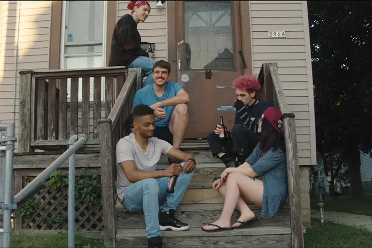 Five people sitting and standing on on a wooden porch in front of a house, holding bottles and talking.