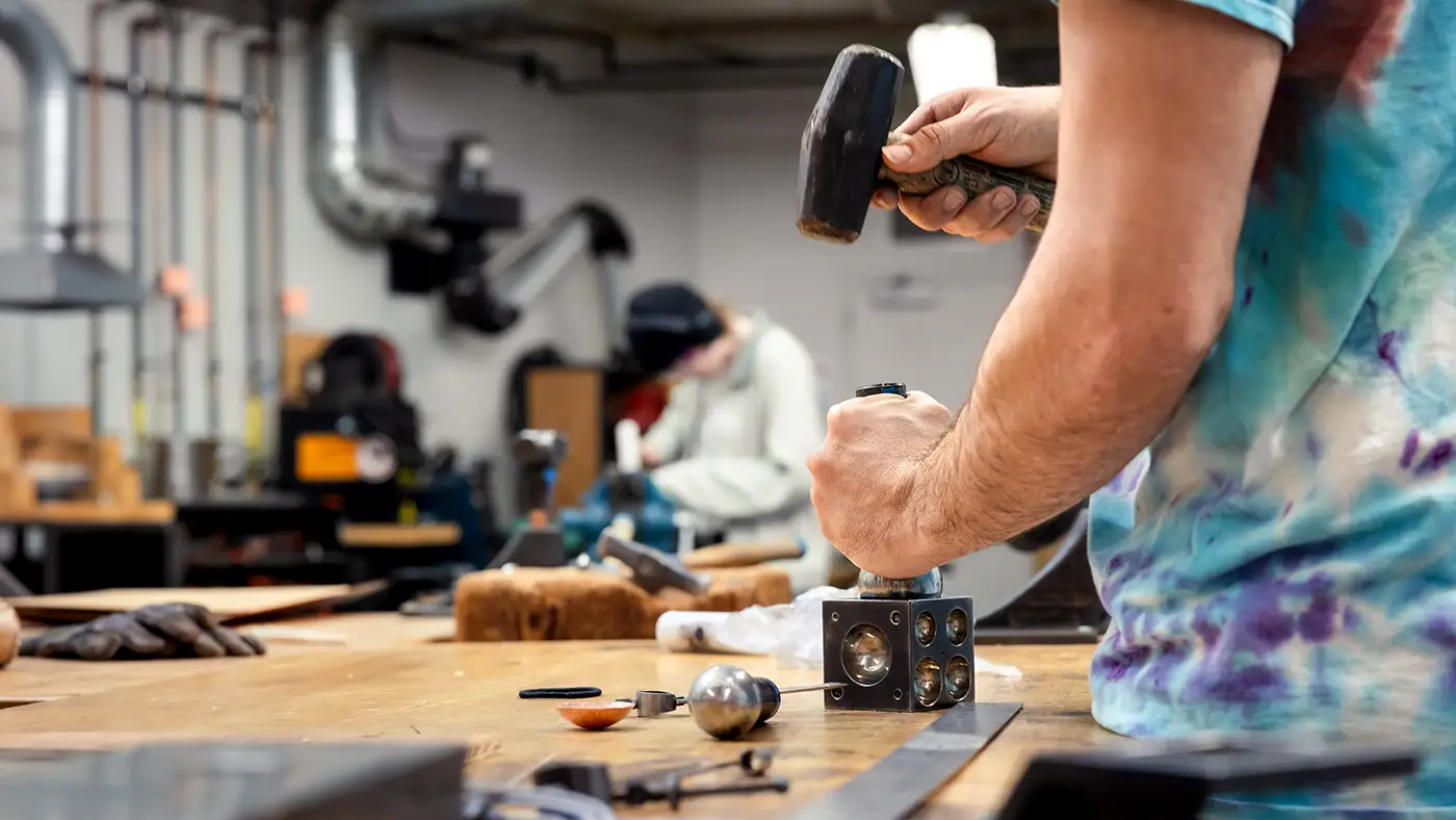 Person using a hammer and metal block at a workshop table with tools scattered around.