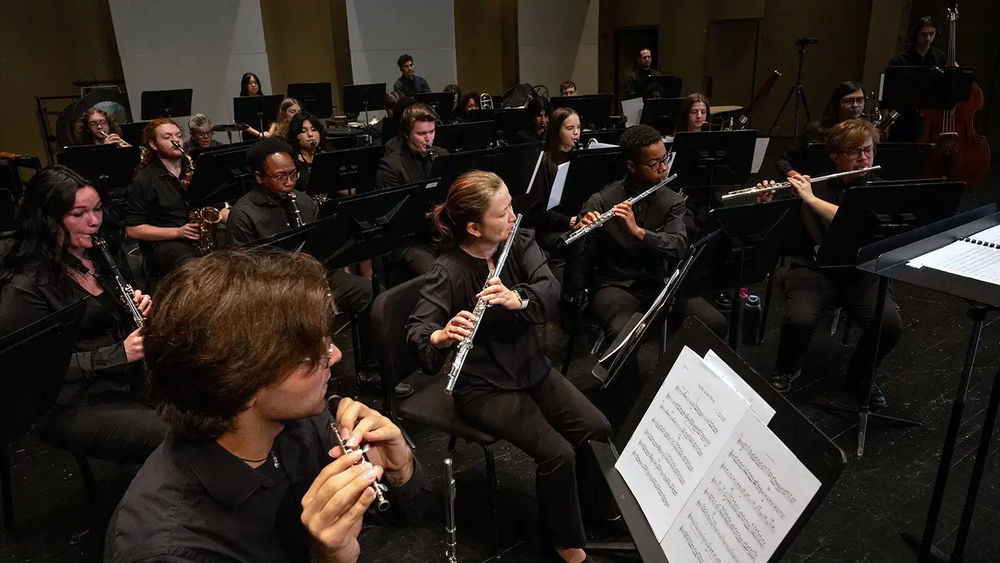 Concert band performing on stage, musicians in black attire playing flutes, clarinets, and saxophones.