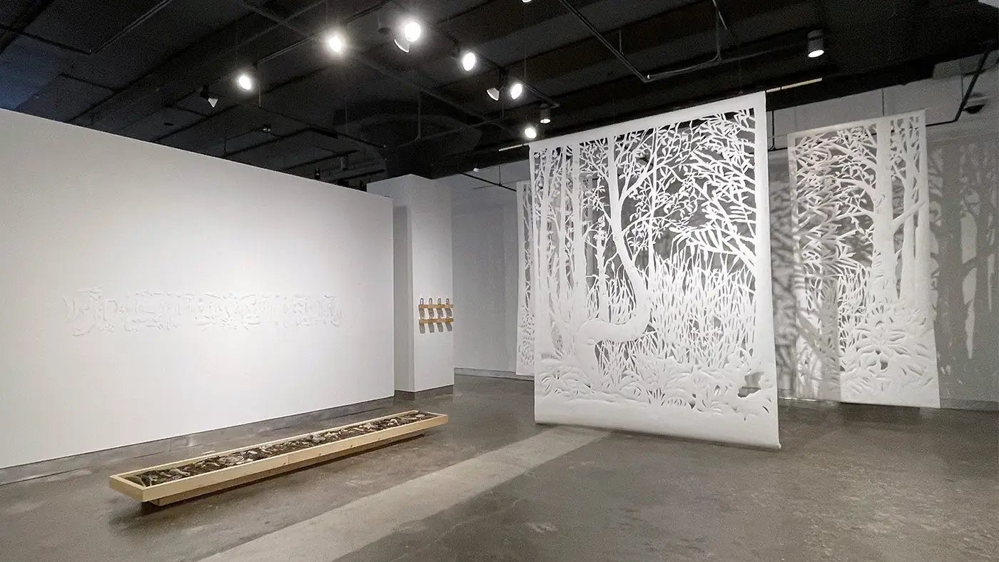 Gallery installation featuring large white paper-cut forest panels suspended in space, casting intricate shadows on surrounding walls.