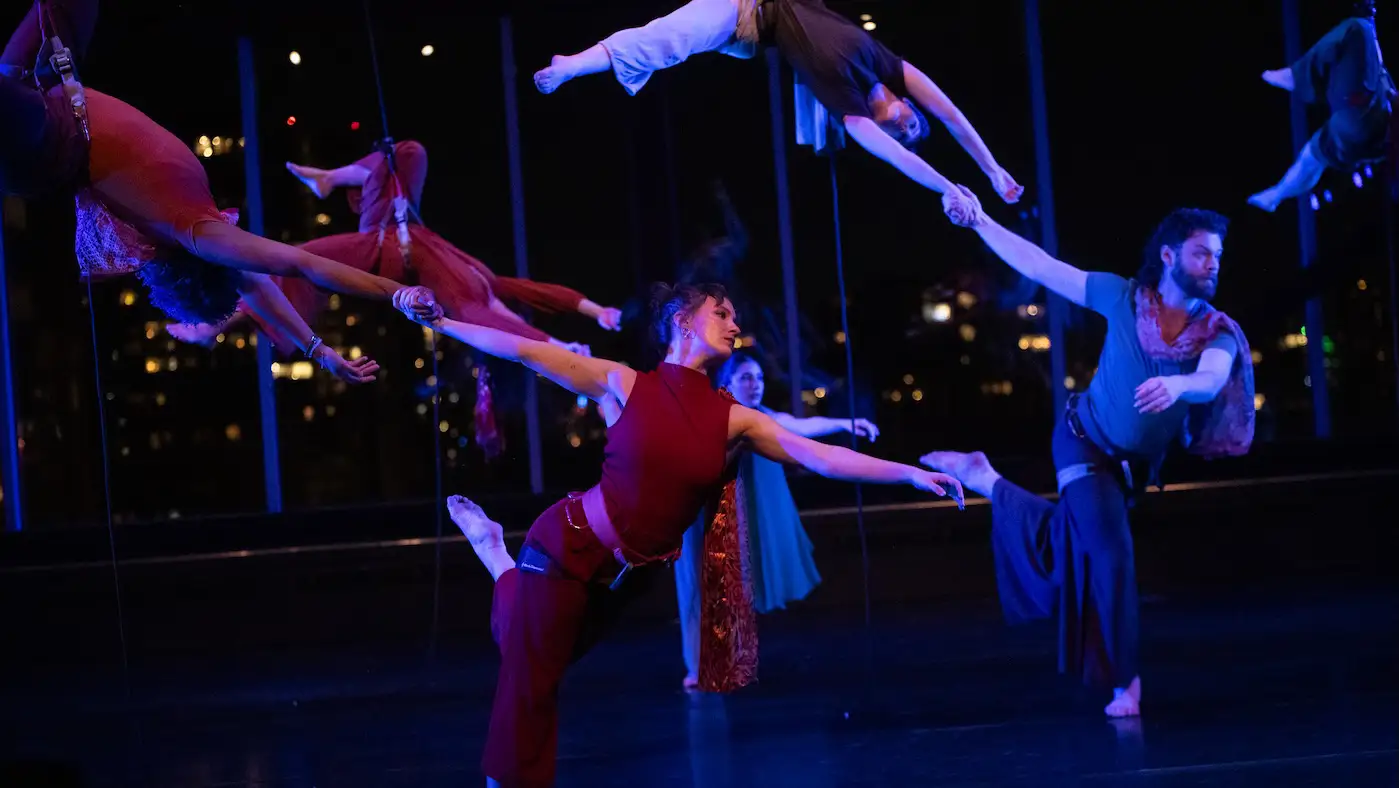 Dancers suspended and grounded move in unison, reaching and balancing under blue stage lighting.