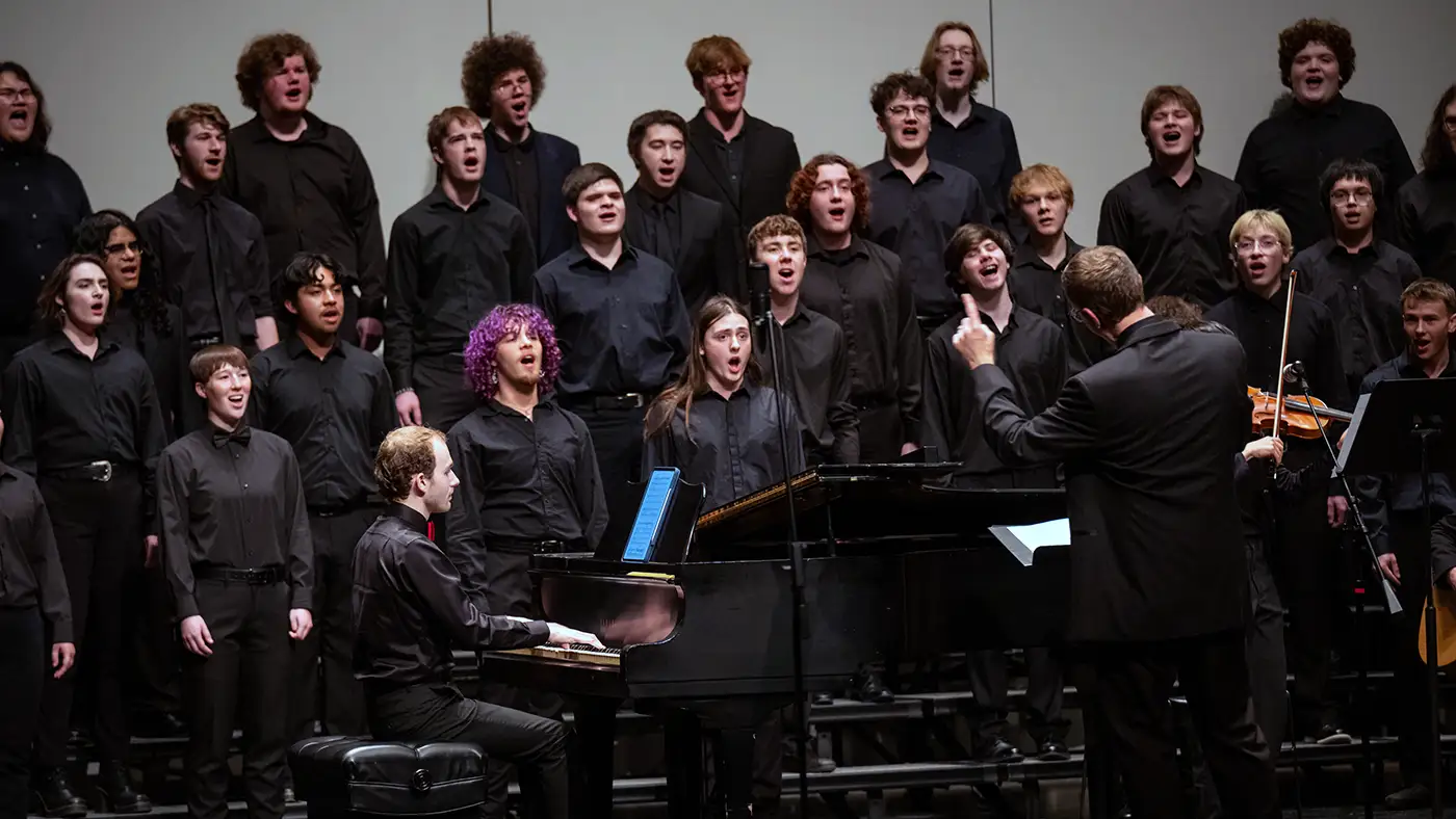 Choir in black on risers sings as conductor leads and pianist plays grand piano on stage.