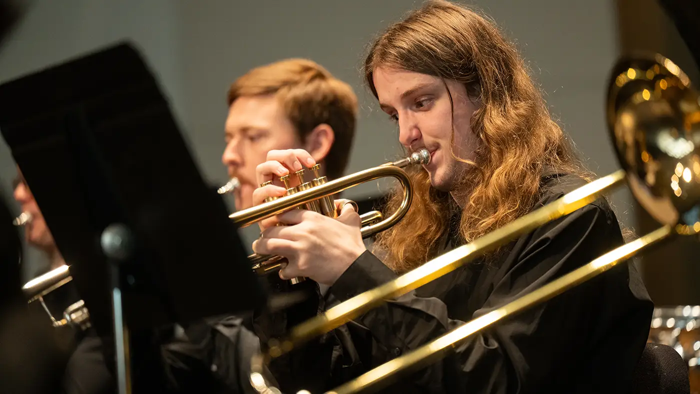 Trumpet player performs in a concert band, brass section visible in foreground and background.