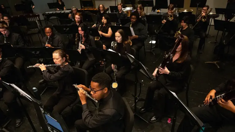 University community band performs on stage, woodwinds and brass seated with percussion behind, all in black attire.