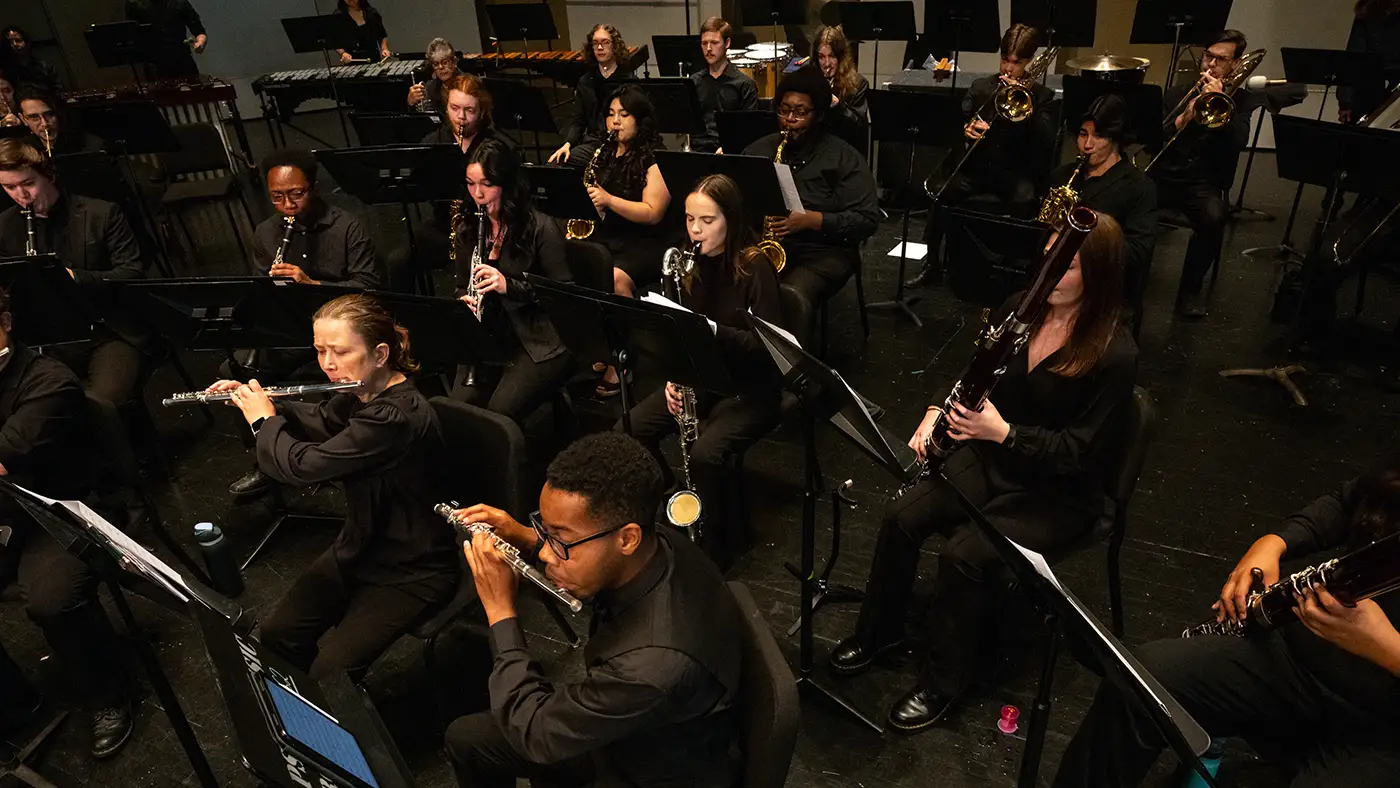 University community band performs on stage, woodwinds and brass seated with percussion behind, all in black attire.