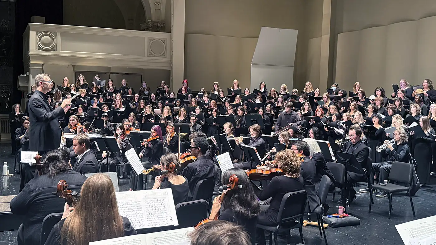 Large orchestra and choir perform on stage with conductor leading ensemble in concert hall.
