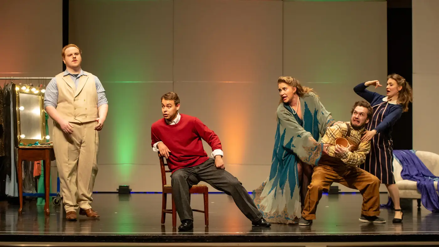Five performers onstage in a lively opera scene, with dramatic poses, a chair, and colorful lighting.