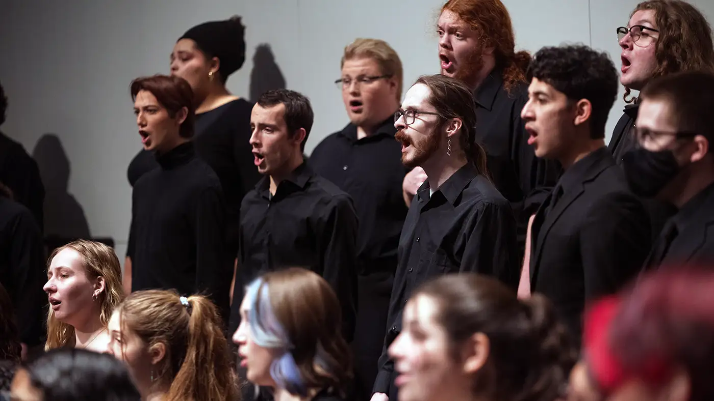 UWM Concert Chorale in black attire singing together on stage in close formation, mid-performance.