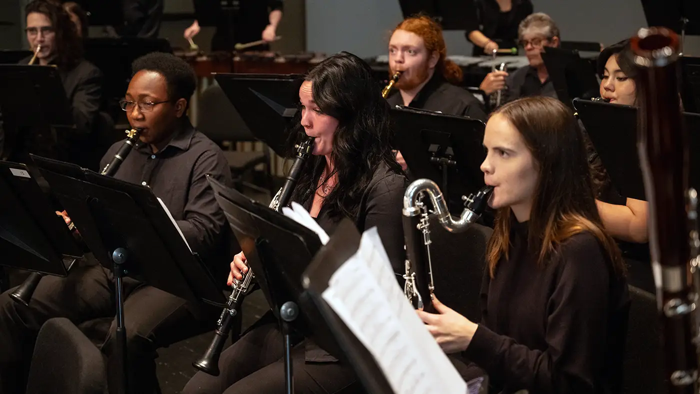 Woodwind musicians in black attire perform on stage, reading sheet music during ensemble concert.