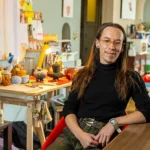 Pavonis sits in their studio leaning on a table.