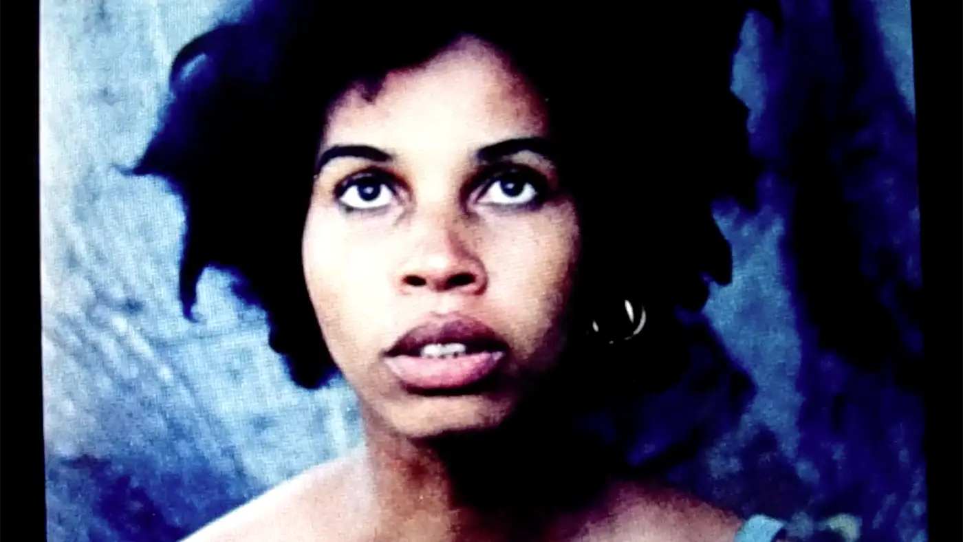 Person with dark, curly hair standing against a dark blue textured background.