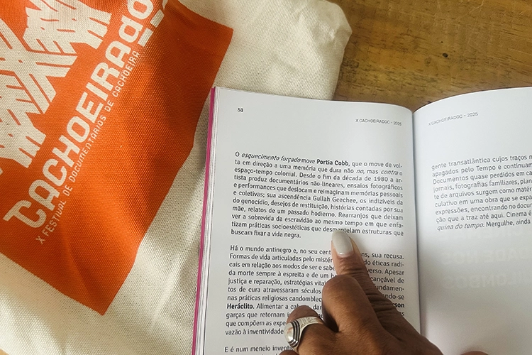 Portia Cobb points to her name in a booklet that references her work in print. A tote bag has a large orange square with the word "Cachoeirado" in white text.