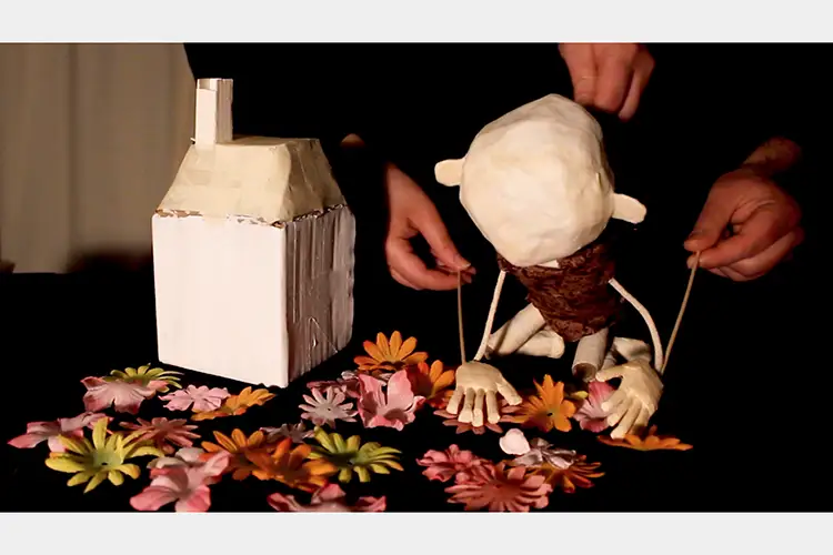 clay puppet of a human on all fours on a table of flowers next to a clay house