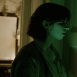 A young woman stands next to a closed door and is lit by green light