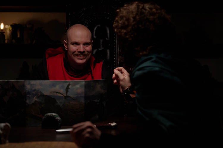 A man smiles nafariously while turing to talk to the man in a green cloack