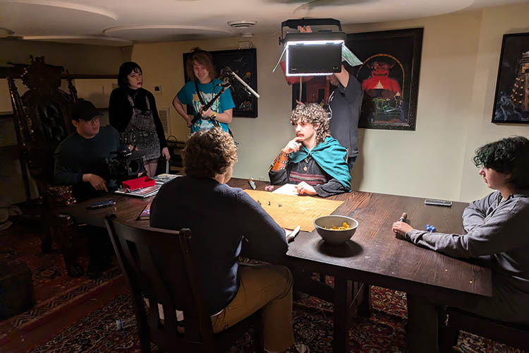 Actors chat around a table while the crew sets up equipment