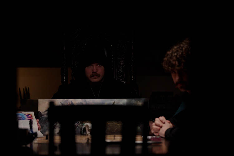 A cloaked man looks to a man dressed in a cape over a privacy screen