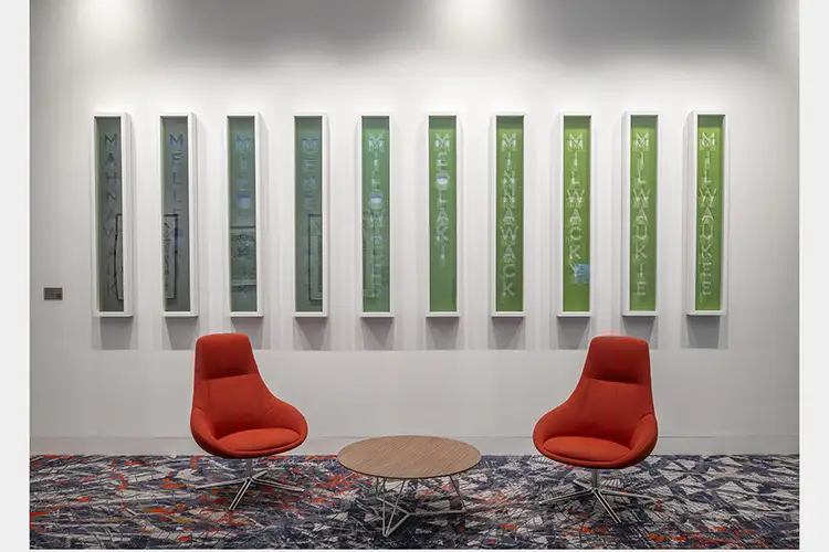 Room with two red chairs on either side of a round coffee table, ten rectangle boxes vertically spell different versions of Milwaukee's names