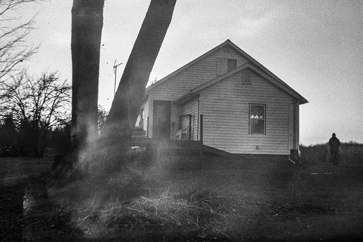 Black and white photo of the side of a house with a large tree in the foreground and a man walking in the background.