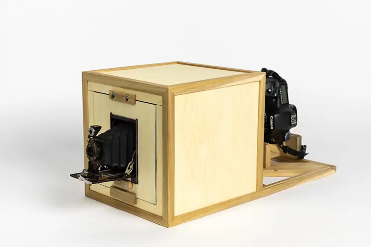 Sculpture of a modern camera leading into an aperture box where an antique camera lens emerges.