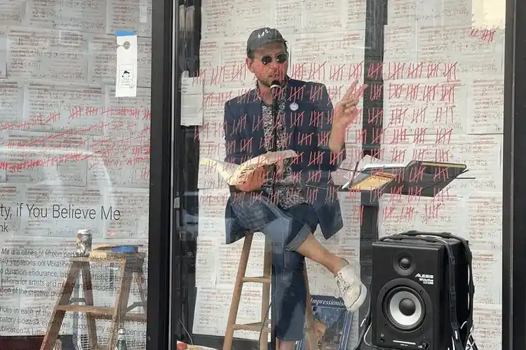 Gif of man reading a book into a microphone inside an outdoor glass room