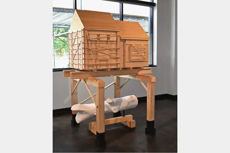 Sculptural boarded up house sitting on wooden frame with plaster cast of a log beneath it