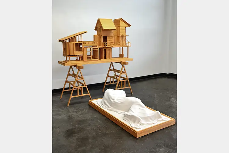 Wood model house sitting on saw horse table with plaster modeled figure at base