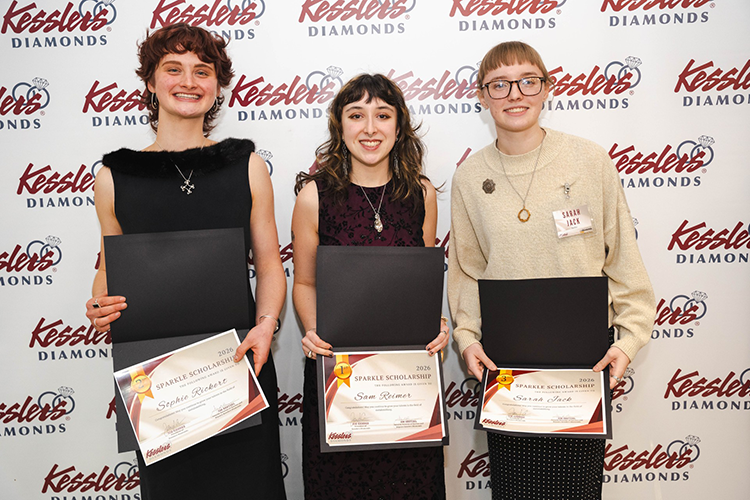 Sophie Rickert (wearing a black dress and a necklace), Sam Reimer (wearing a dark red dress and a necklace), and Sarah Jack (wearing glasses, a black maxi skirt, and a white sweater) smile while holding their scholarships. The stand before a backdrop that says "Kesslers" in a red, cursive font.