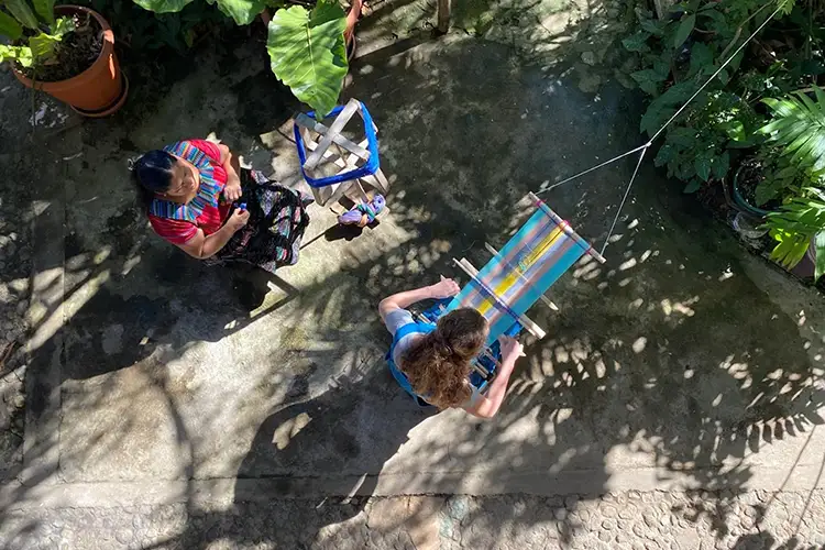 As seen from above, Liz Rex weaving a cloth out of blue, pink, white, and yellow while another woman looks on