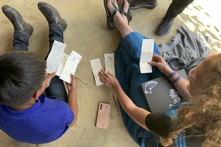 As seen from above, Liz Rex working with a young child, both sitting on the floor, holding different paper drawings of monsters