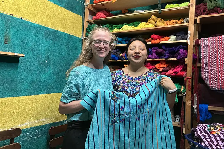 Liz Rex and another woman standing in front of a shelf containing many colorful yarn bundles, they are holding a bright blue, striped cloth