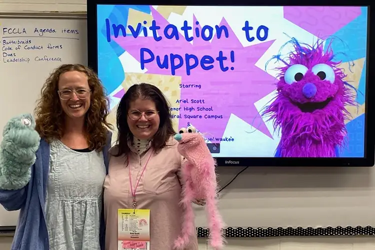 Liz Rex standing with a student, hold holding a hand-puppet each, in front of a screen showing a presentation titled "Invitation to Puppet"