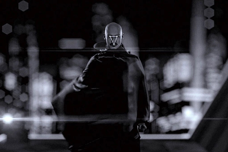 A bald man wearing a cloak stands up against a cityscape. The photo is black and white. There is a white triangle.