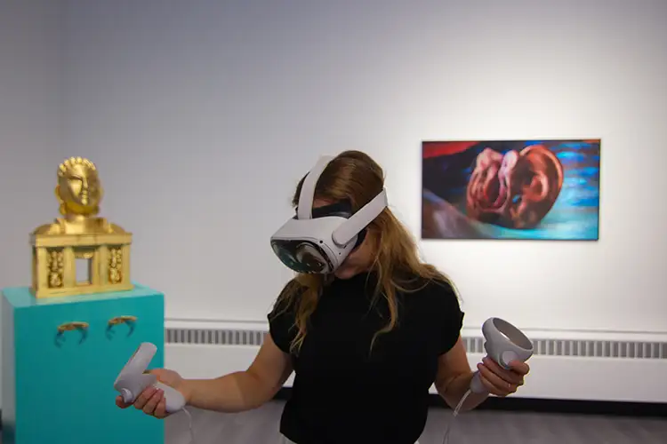 A woman wearing a virtual reality headset, looking down. She stands in front of other various art pieces. A woman wearing a virtual reality headset, looking down. She stands in front of other various art pieces.