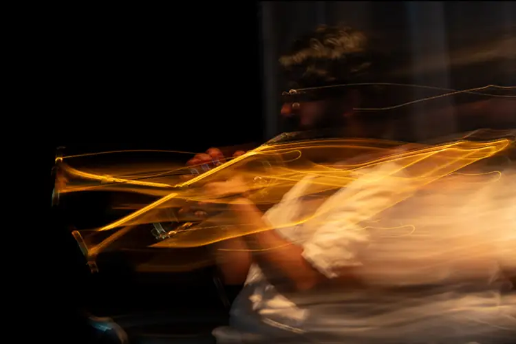Motion-blurred image of a person playing a trumpet, with golden light trails tracing the instrument and hands against a dark background.