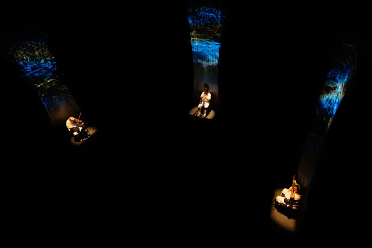 Dark gallery installation showing three seated performers in separate pools of light beneath tall, vertical projections glowing blue and gold.