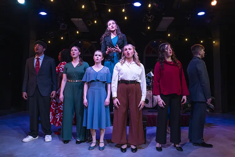Group of performers standing on a stage under lights, singing as an ensemble.