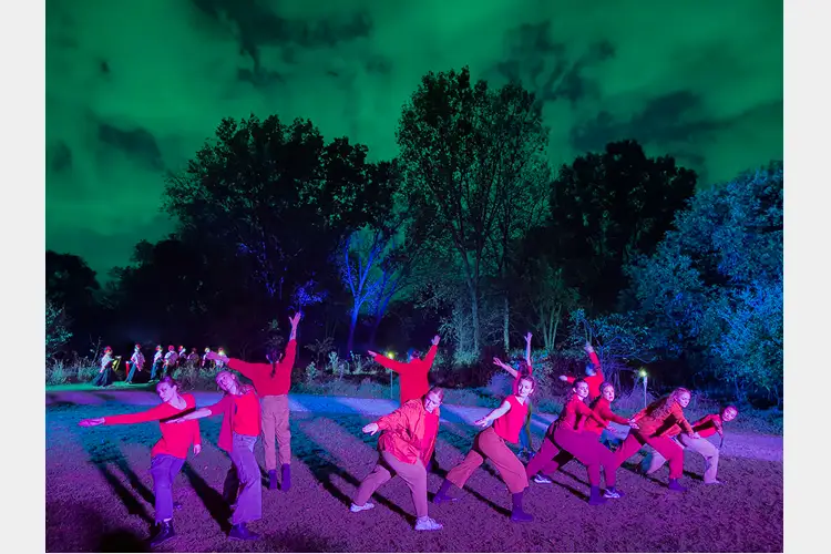Group of performers in red outfits pose mid-movement outdoors at night, lit with vivid blue and purple lighting against dark trees and sky. Group of performers in red outfits pose mid-movement outdoors at night, lit with vivid blue and purple lighting against dark trees and sky.