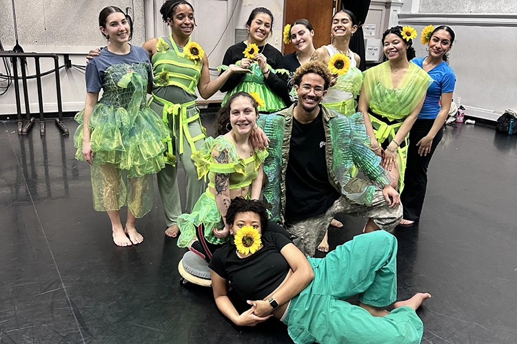 Picture of the Springdances cast. They're wearing outfits in different shades of green. They smile towards the camera, with the first most person holding a sunflower in their mouth.