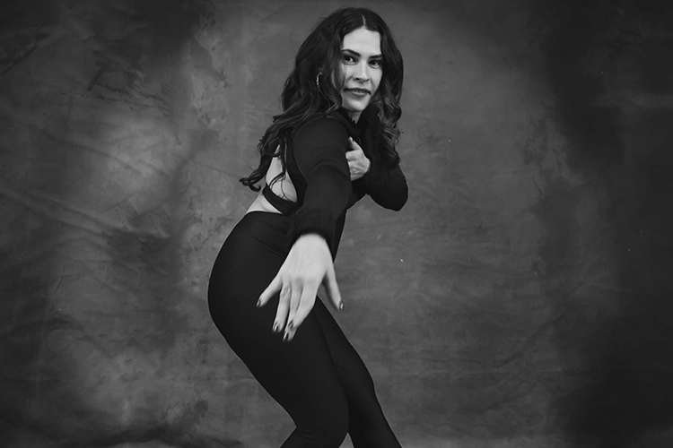 Karlies Kelley in black outfit reaching forward in a dynamic pose against textured studio backdrop.