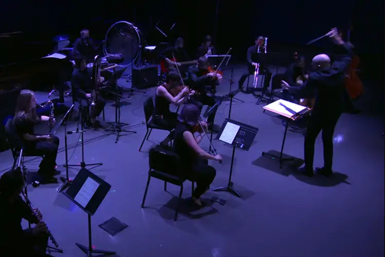 Wide view of a contemporary ensemble performing onstage, with a conductor leading string, wind, and percussion players seated under cool blue stage lighting.
