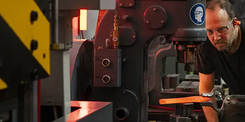 Two people observe a glowing metal press shaping hot steel in a blacksmithing studio.