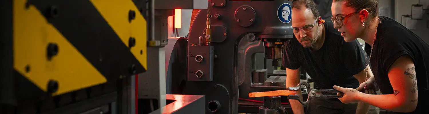 Two people observe a glowing metal press shaping hot steel in a blacksmithing studio.
