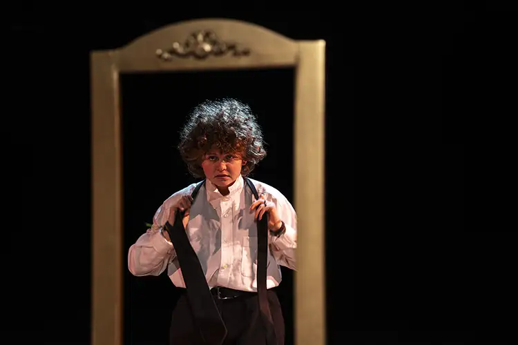 Performer on stage, framed by a mirror, adjusting suspenders under spotlight.