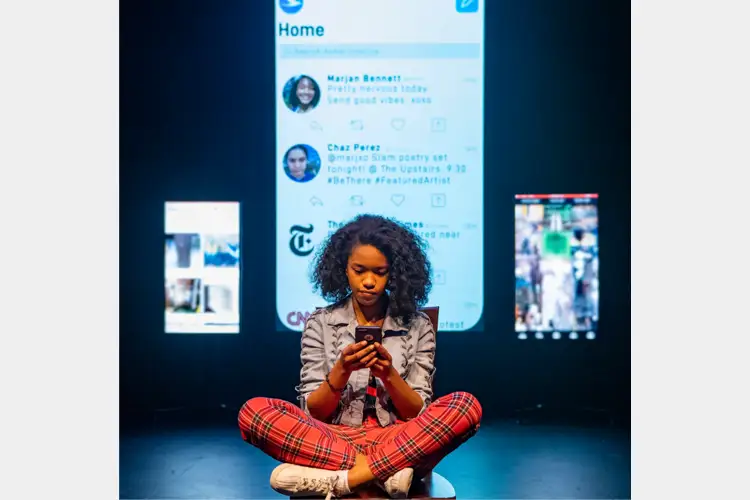 Person sits cross-legged on stage, looking at a phone, with a large projected social media feed displayed behind and screens on both sides. Person sits cross-legged on stage, looking at a phone, with a large projected social media feed displayed behind and screens on both sides.