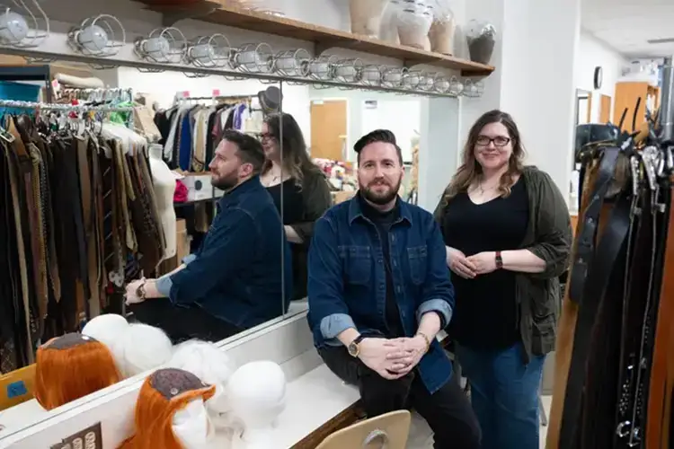 Orlenko and Rochester smile and pose in the costume shop. They stand in front of a mirror surrounded by wings and costumes.